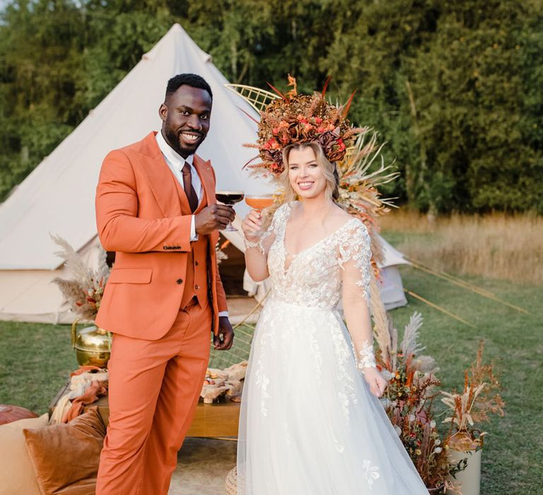 Bride in bold autumnal headpiece with brown & orange silk flowers and pheasant feather details and long sleeved v neck soft ivory dress having a cocktail with groom in burnt orange rust coloured suit with brown tie, pocket square, brown accessories and dried flower, rose and pampas grass boutonniere  