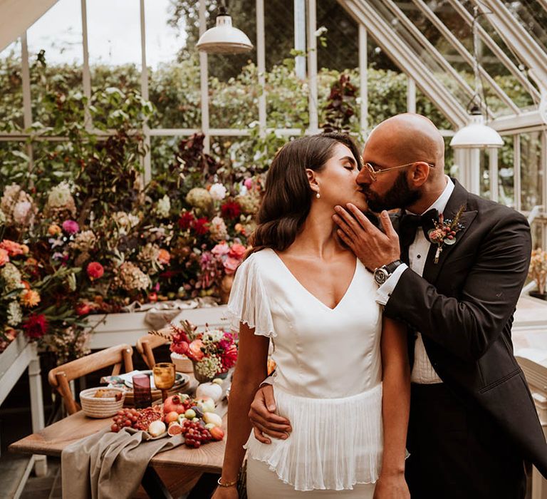 Groom in classic black tuxedo with autumnal dried flower boutonniere and black bowtie kissing bride in v-neck satin wedding dress with sheer butterfly sleeves in glasshouse wedding venue