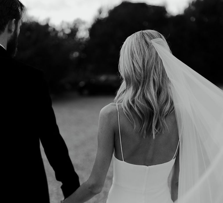 Bride in elegant fitted wedding dress with thin spaghetti straps walking holding hands with the groom for minimal style wedding 