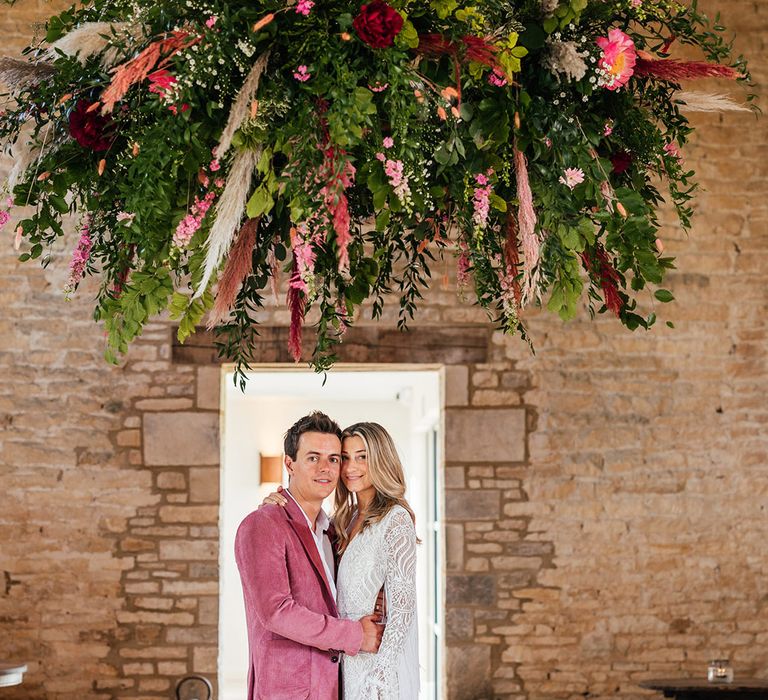 Bride and groom embracing under suspended foliage, rose, pampas grass and eucalyptus decor at Old Gore Barn wedding venue