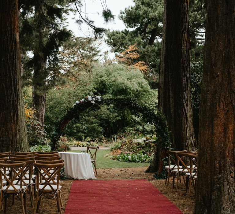 Red carpet leading to green foliage archway with wooden chairs lining the aisle for outdoor ceremony at the Royal Botanic Garden Edinburgh