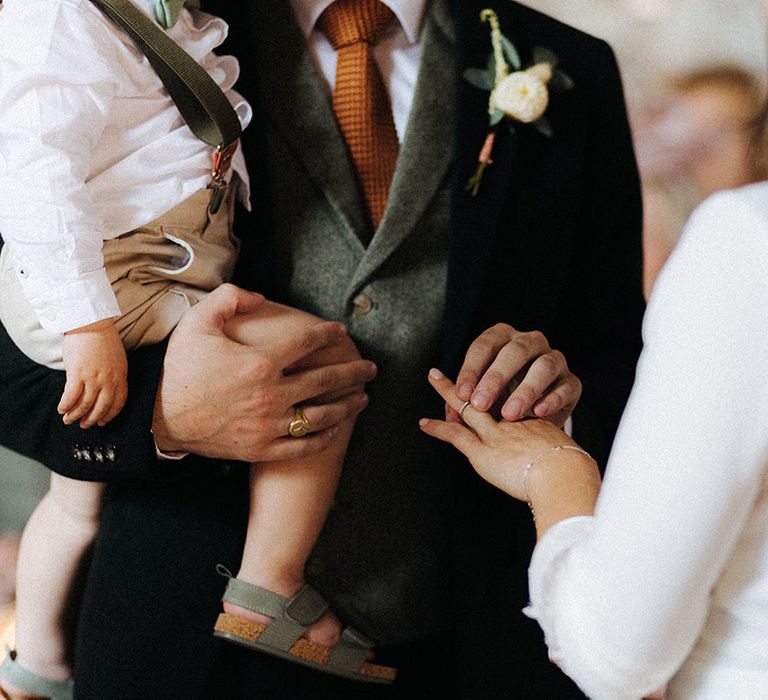 Groom in a three piece suit holding the toddler puts the bride's wedding band on 