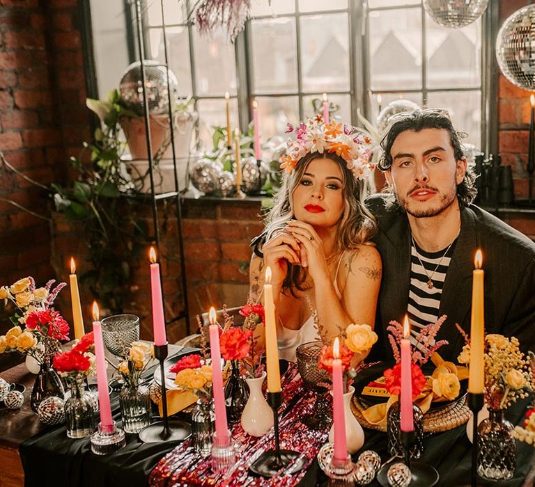 Bride in satin v-neck wedding dress and artistic floral crown and groom in black and white long sleeve shirt and grey pinstripe blazer sitting at colourful wedding tablescape with pink metallic streamers, hanging disco ball decorations, and tapered candles 