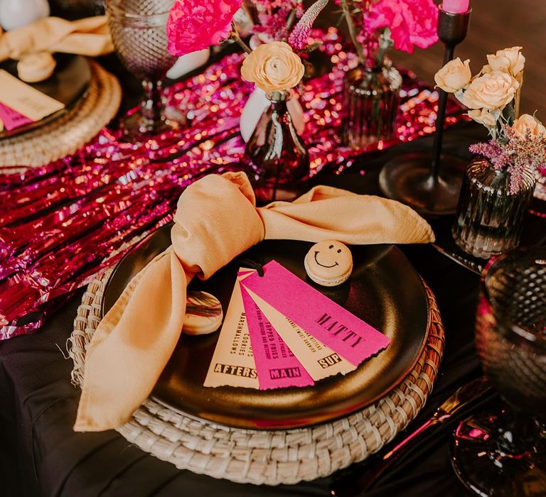 Colourful wedding tablescape with pink metallic streamers, pink rose and yellow peony decorations, pink and yellow tapered candles, yellow napkins, smiley face decorations and hot pink and yellow place names 