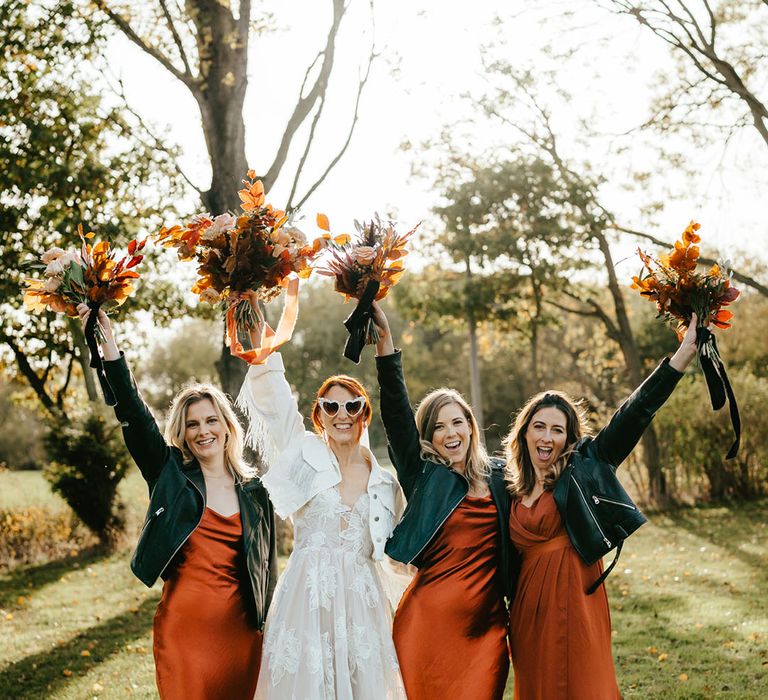 Bride stands beside her bridesmaids in burnt orange satin bridesmaid dresses and black leather jackets 