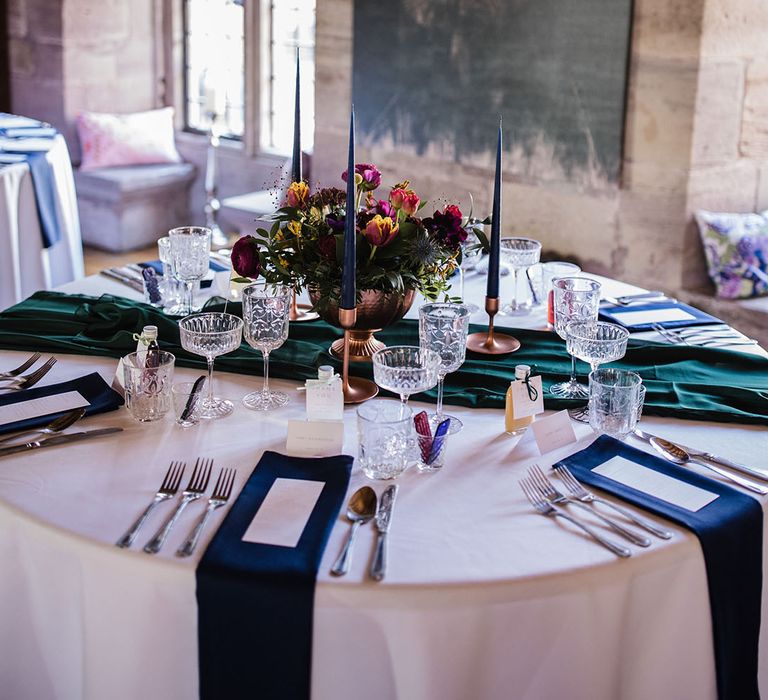 Round tables with white tablecloths with emerald green table runner and blue napkins to match the blue taper candles with crystal glassware and a spring flower table centrepiece