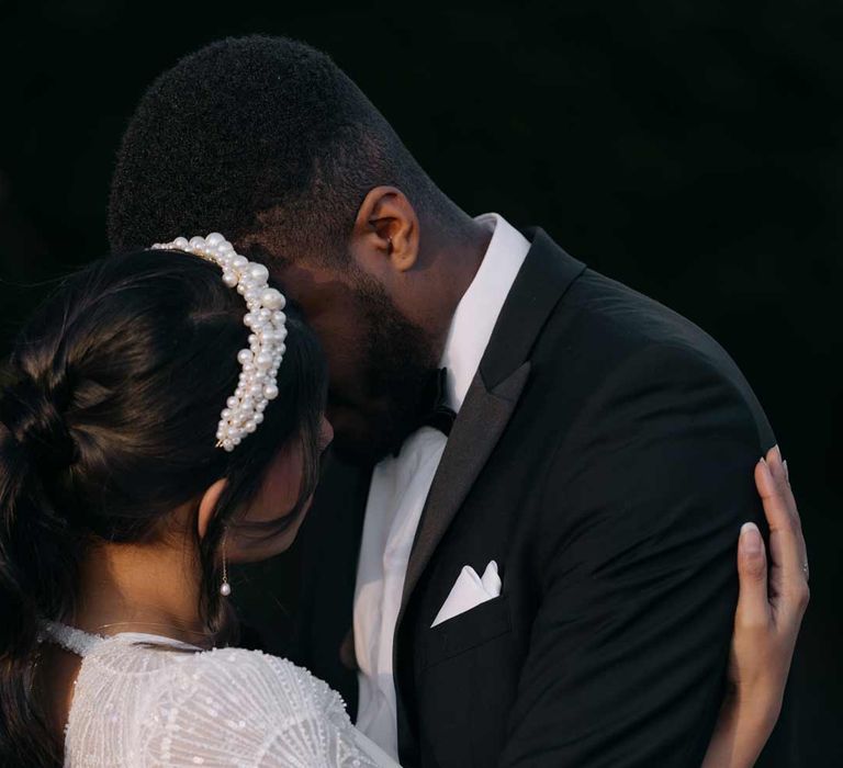 Groom in classic black tuxedo, black bowtie and white pocket square embracing bride in sparkly lace detail open back wedding dress and large pearl headband 