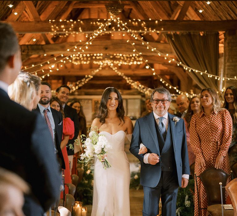 Father of the bride in a navy blue suit walking the bride down the aisle in a strapless wedding dress with detachable pearl sleeves for rustic barn wedding