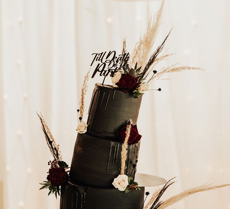 Black wedding cake with three tiers and drip icing effect decorated with dark red and white rose flowers with pampas grass and black cake topper