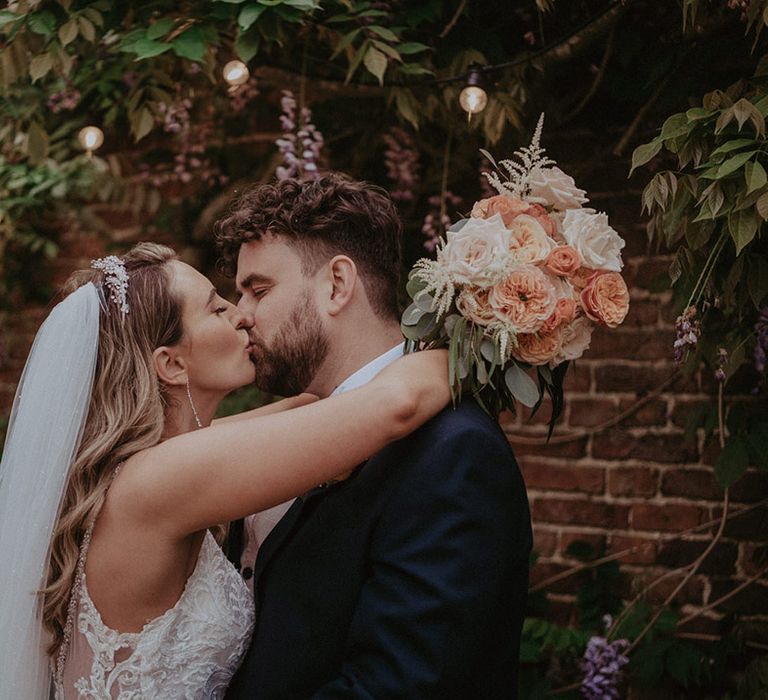 A rustic luxe Stanlake Park wedding with the bride in a fitted lace wedding dress with tiara and veil kissing the groom in a dark suit jacket 