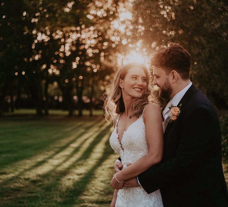 The bride in an embellished lace bridal gown is embraced from behind by the groom during golden hour 