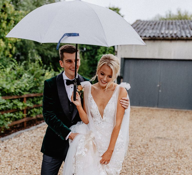 The bride and groom walk together under a large umbrella to avoid the rain on their wedding day 