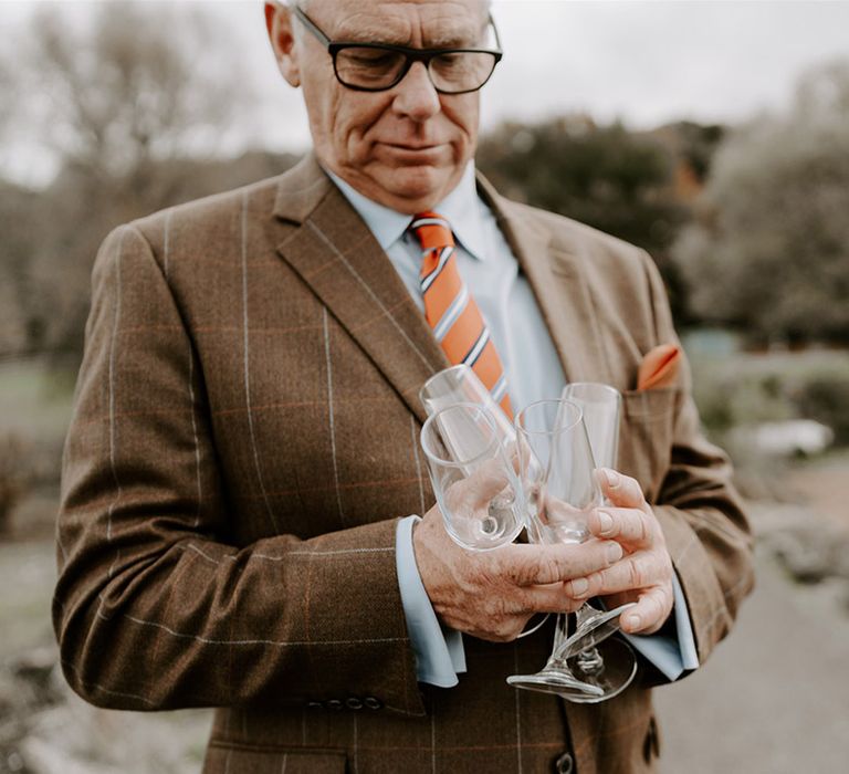 Wedding guest in brown suit with orange checkered detail for autumnal wedding outfit holding champagne glasses 