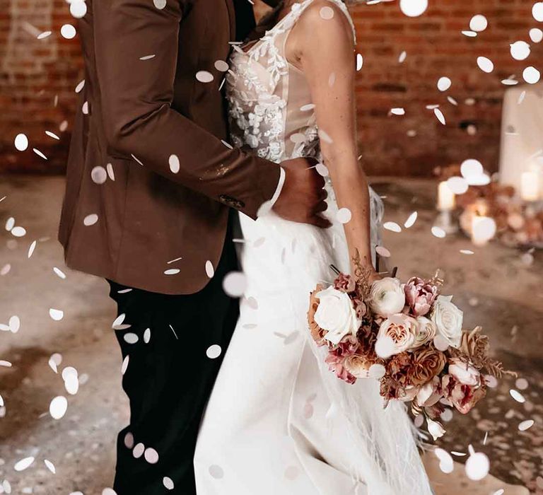 Bride in  corset top sheer floral 3d appliqué wedding dress with fringe overlay and groom in mocha brown blazer, black waistcoat, polka dot tie and pocket square and dried blush boutonniere with white wedding confetti falling around them 