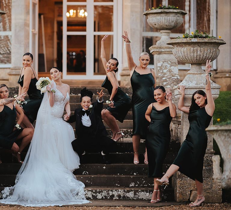 Bridesmaids in midi length black bridesmaid dresses posing with brides in lace sleeveless v neck Enzoani wedding dress with tulle finish and church length veil and black snakeskin print tuxedo with black bowtie and white rose boutonniere at Grittleton House