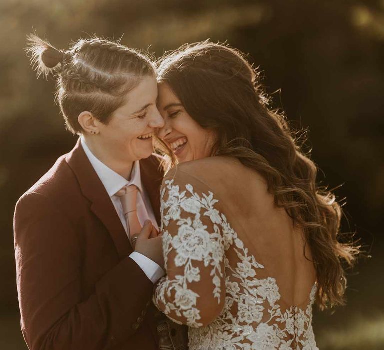 Bride with low back detail long sleeve lace illusion sleeve wedding dress with overlay embracing bride in maroon bridal suit with grey waistcoat, dusky pink tie and autumnal dried flower boutonniere at golden hour wedding photoshoot at Bassmead Manor Barns 