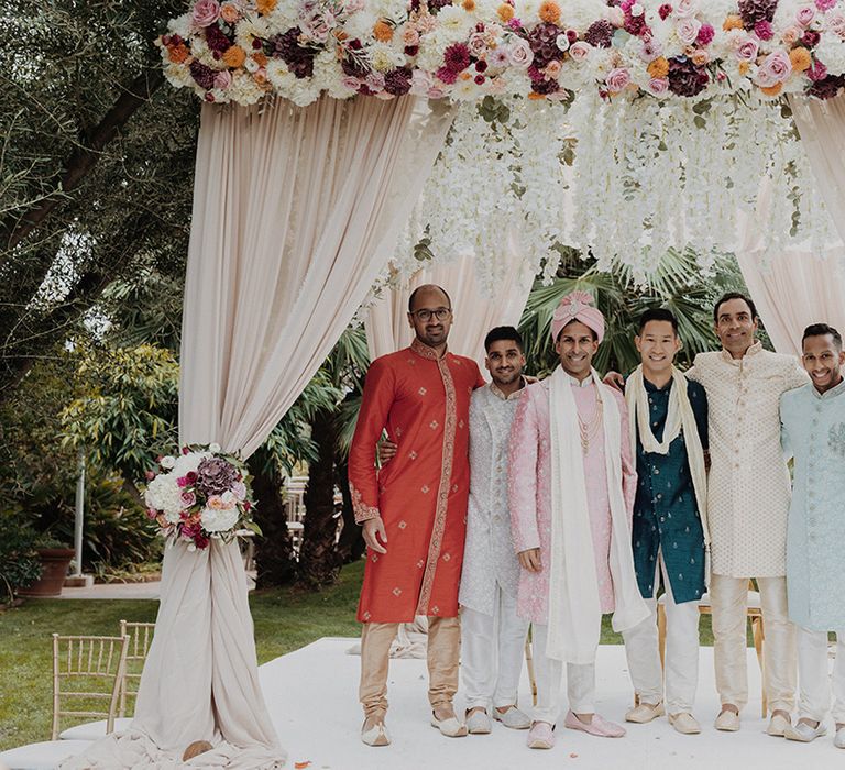 Groomsmen in the wedding mandap wearing mismatched grooms sherwanis and groom wearing light pink sherwani, light pink turban and cream trousers  