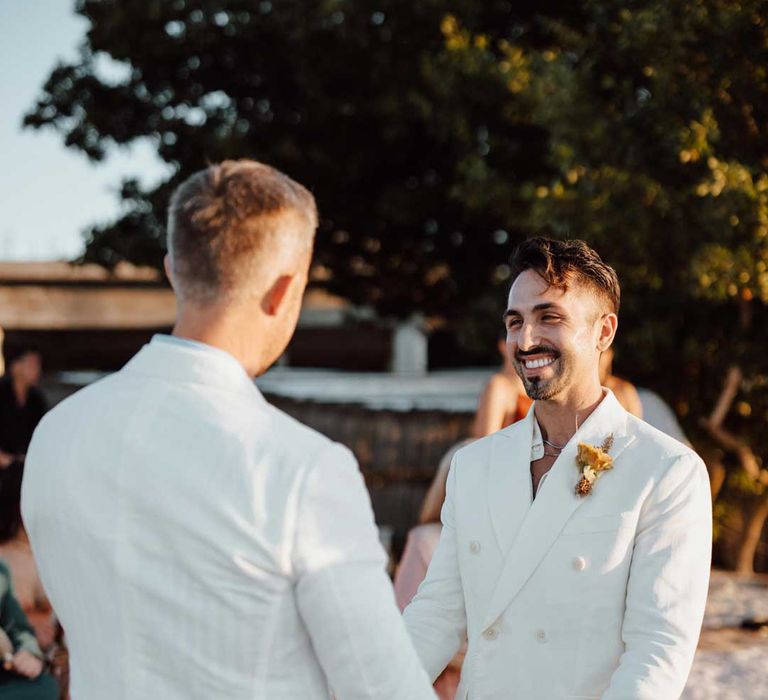 Groom in mens white wedding suit, dusky orange garden rose and dried flower boutonniere, white shirt, and silver jewellery holding hands with groom in white double breasted suit 