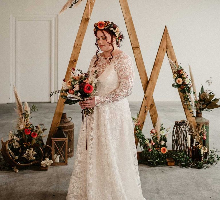 Bride in 3D appliqué long sleeve wedding dress standing in front of one small and one large wooden triangular wedding arch with pampas grass, foliage, and garden rose arrangements attached, rustic wedding decorations, large plants in vases and pillar candles at The Barn at Avington