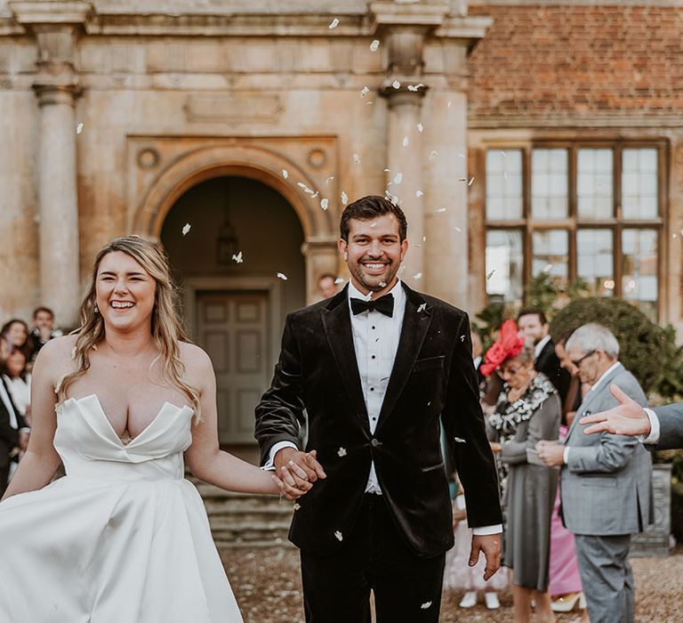 Bride in strapless wedding dress by Suzanne Neville walking hand in hand with the groom in back tie to confetti 