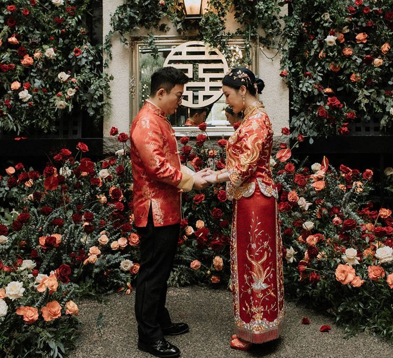 The bride and groom hold hands at their wedding ceremony in red and gold wedding attire for their tea ceremony 