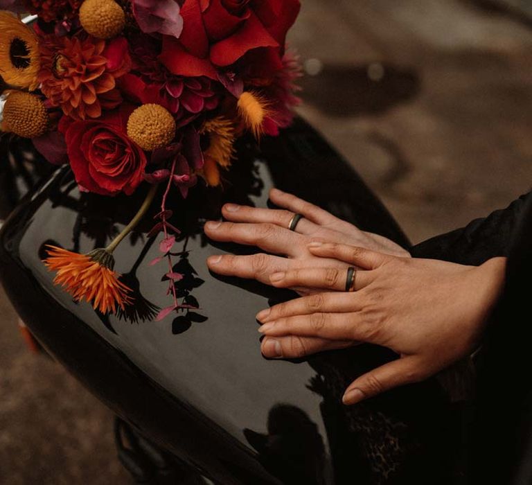 Bride and groom wearing black wedding rings on the back of a motorbike with red vibrant flower arrangements