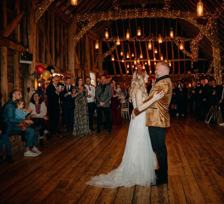 Bride in off shoulder lace wedding dress with corset style top and red rose and gold halo bridal crown dancing with groom in crushed velvet burnt orange grooms blazer and dried flower boutonniere for their first wedding dance at Southlands Barn