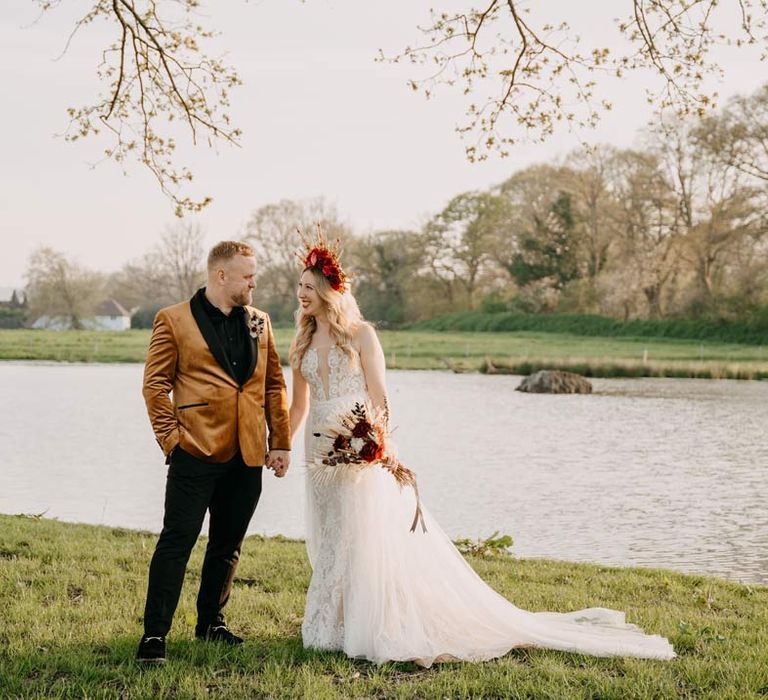 Groom in crushed velvet burnt orange grooms blazer and dried flower boutonniere holding hands with bride in off shoulder lace wedding dress with corset style top and red rose and gold halo bridal crown holding red rose, pampas grass and dried flower bridal bouquet