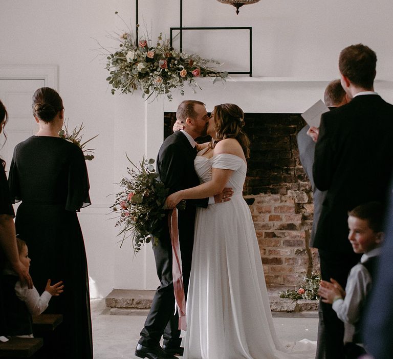 Bride in off the shoulder wedding gown kissing groom in black suit for the first time as a married couple