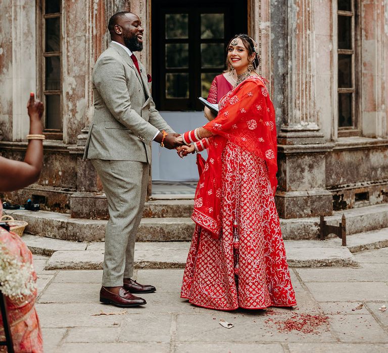 Bride in red and gold wedding lehenga and groom in stylish light grey double breasted suit jacket holding hands 