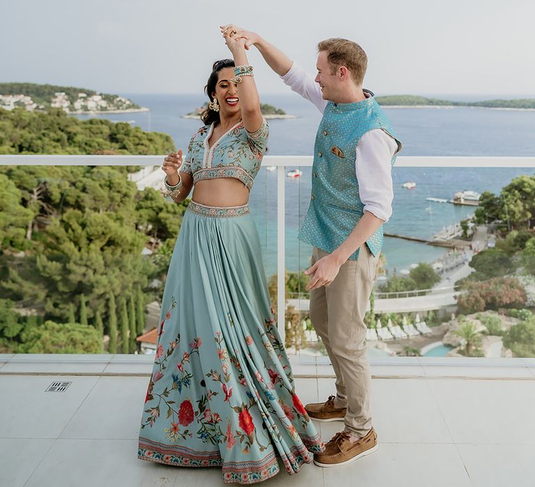 Bride and groom in traditional turqouise South Asian outfits dance in front of ocean view