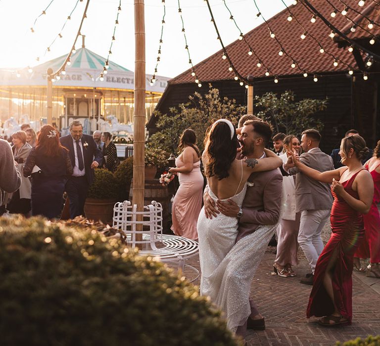 Groom in pink wedding suit bending the bride back in a dance by carousel 