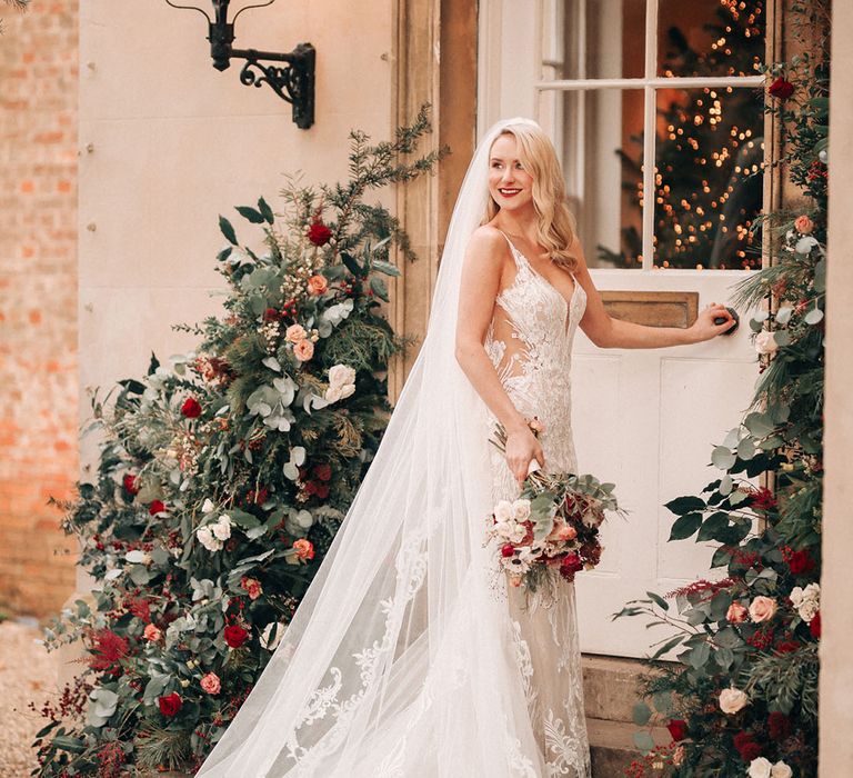 Bride in embellished wedding dress with delicate embroidered veil walking into venue with Christmas tree decor and flower columns 