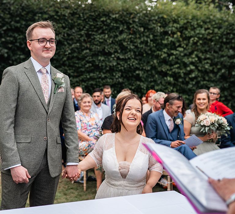 Bride in lace wedding dress from BRIDE by Aster with the groom in a grey wedding suit at their outdoor wedding ceremony 