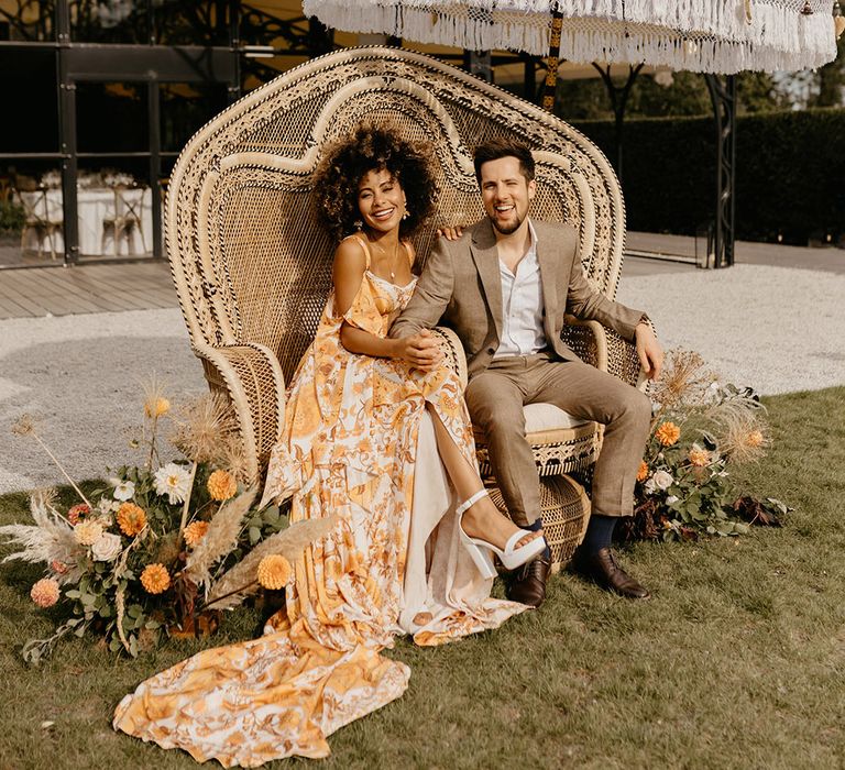 Bride in yellow orange floral patterned wedding dress with peacock chair sitting with groom under fabric tassel umbrella 