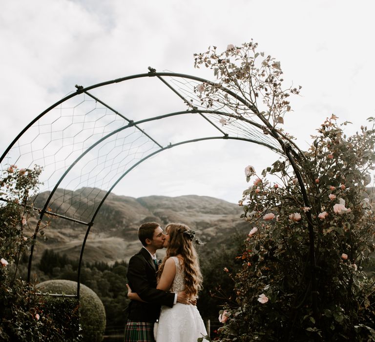 Bride and groom share a kiss next to some rose bushes in their traditional dress 