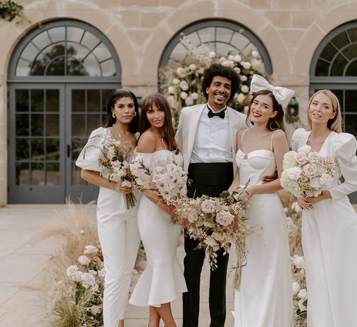 Wedding party portrait white groom in a white tuxedo jacket and the bridal party all in white dresses and jumpsuits.