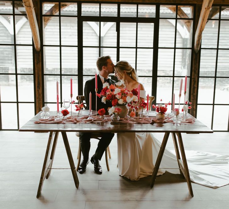 Bride & groom sit in front of Barn window lined in black with coral themed tablescape in front of them