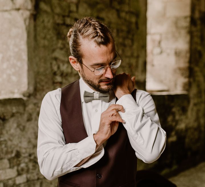 Groom in a burgundy suit with waistcoat and bow tie adjusting his cufflinks