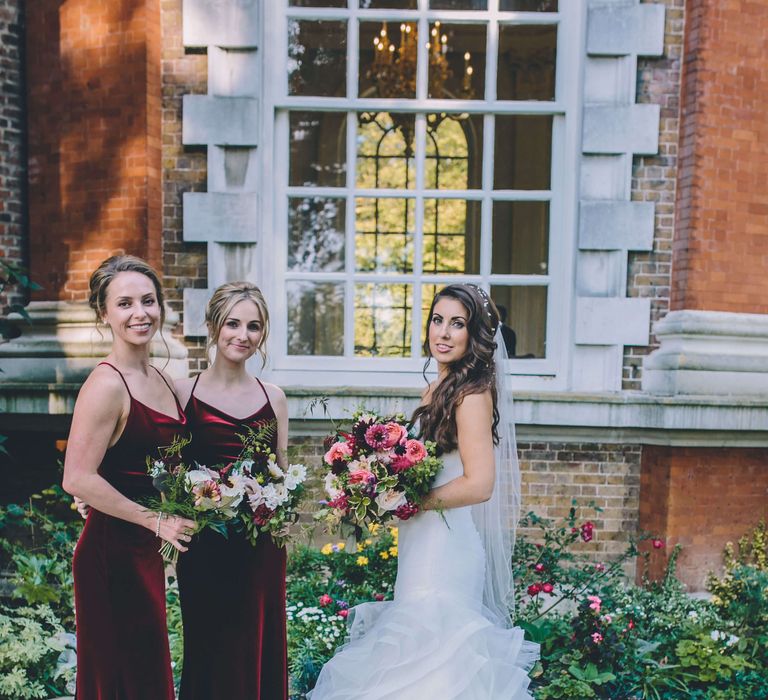 Bride with two bridesmaids outside London wedding venue, bridesmaids wear deep red velvet bridesmaid dresses with pink rose bridal bouquet