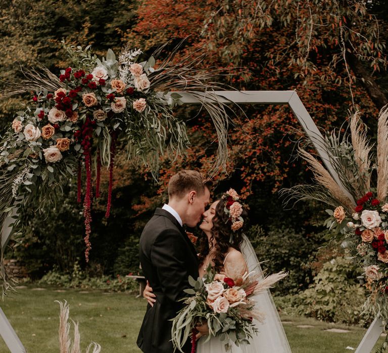 Groom in a tuxedo kissing his bride in a tulle skirt wedding dress with flower crown and oversized bouquet 