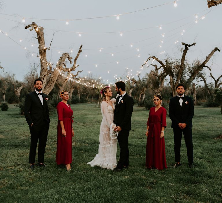 Bride in long sleeved boho wedding dress holding hands with groom in navy suit with bow tie standing in olive grove lit up with fairy lights next to bridesmaids in fuchsia dresses and groomsmen in suits and bow ties at enchanted forest wedding in Italy