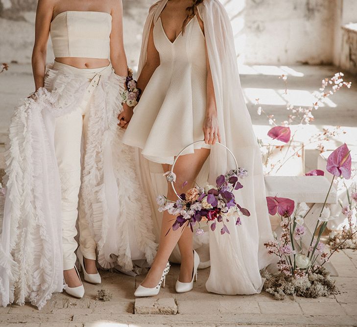 Bride in a mini wedding dress with bridal cape holding a hoop bouquet and bride in trouser and crop top separates with detachable ruffle skirt surrounded by purple flowers