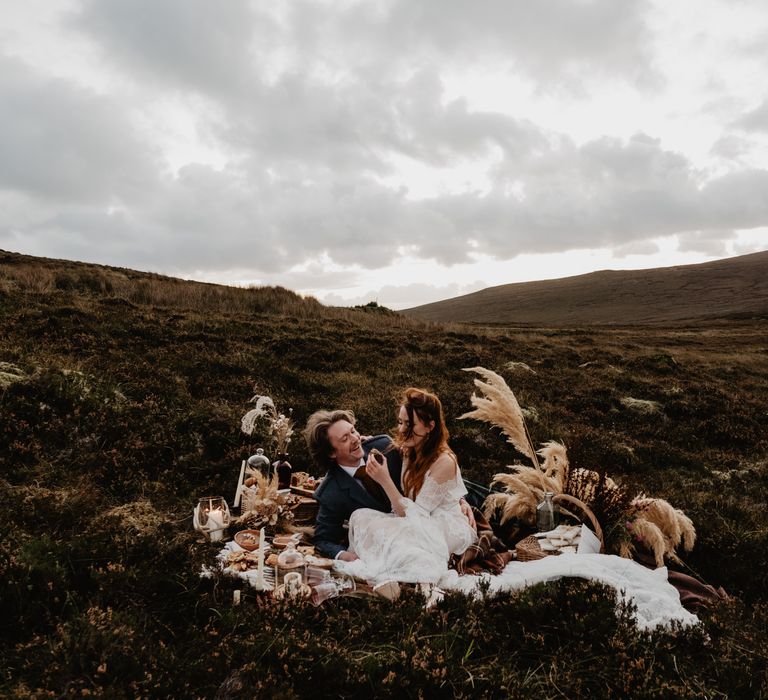 Bride & groom sit down to picnic on the hillside with pampas grass decor