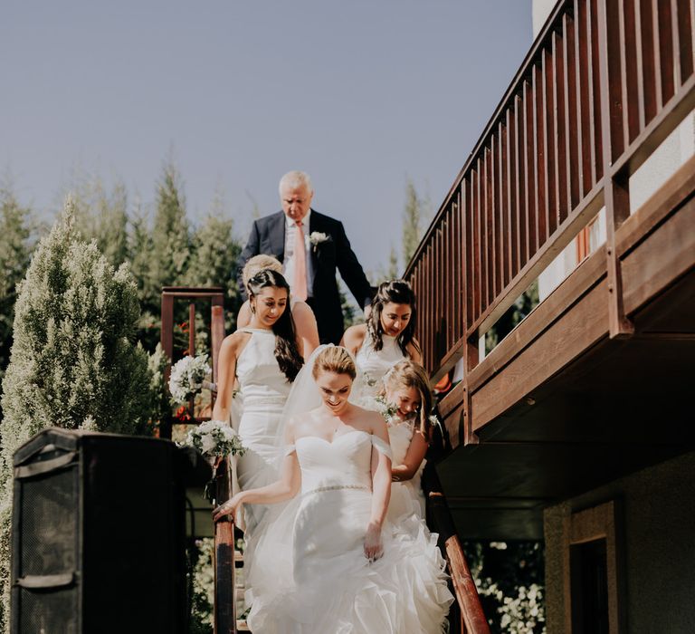 Bride descending the stairs wearing an off the shoulder ruffle skirt gown, followed by her bridesmaids and father