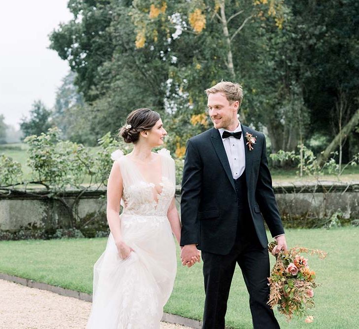 Groom in a tuxedo and horse waistcoat holding hands with his elegant bride in a tulle wedding dress and pinned bridal updo