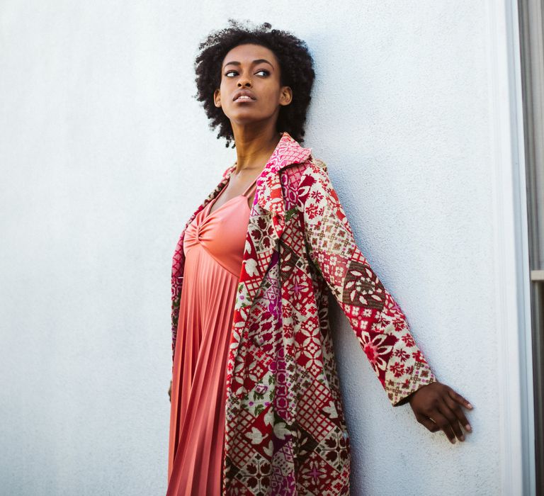 LGBTQ+ Bride-to be in a coral dress and patterned long-line coat standing next to a white wall at engagement photoshoot 