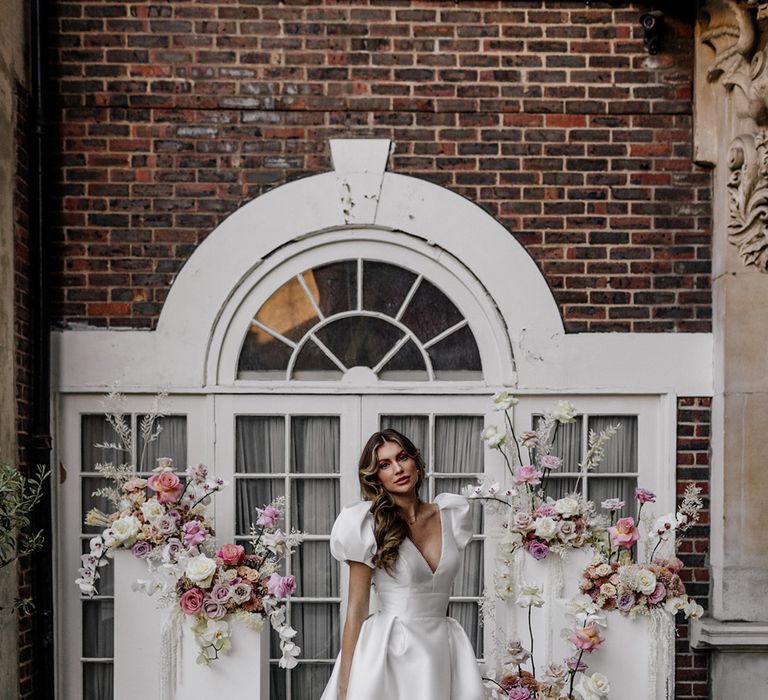 Bride in a stylish Love In Lace Bridal gown with puff sleeves and front split standing next to blush pink floral arrangements 