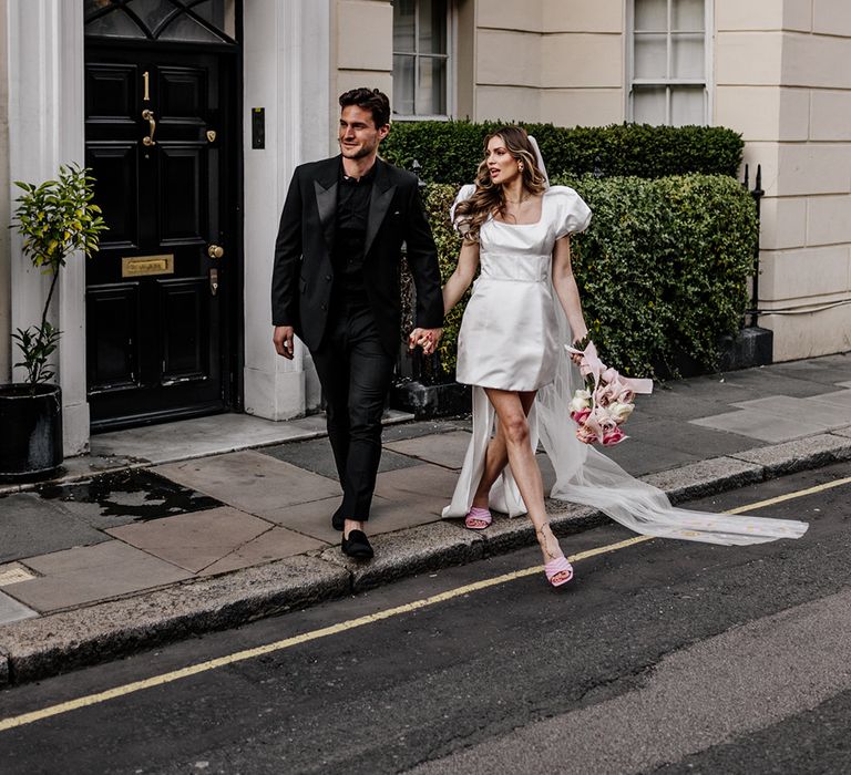 Groom in a black shirt, suit and loafers crossing the road with his bride in a short wedding dress with puff shoulder detail 