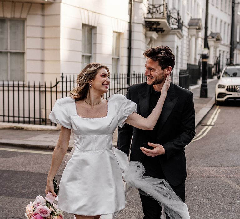 Bride in a short wedding dress wearing pink mules and holding a blush, peeled back rose bouquet embracing her grooms face 